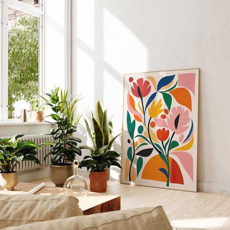 May include: A framed floral art print with a colorful abstract design. The artwork features pink, red, yellow, and blue flowers and leaves against a cream background. The print is displayed in a bright, sunlit room with potted plants.