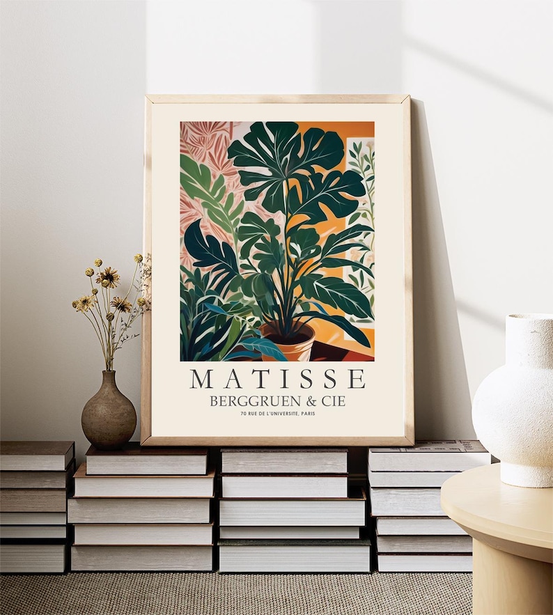 Puede incluir: Una impresi&oacute;n art&iacute;stica enmarcada con una ilustraci&oacute;n bot&aacute;nica de una planta en maceta con grandes hojas verdes. La impresi&oacute;n incluye el texto "MATISSE BERGGRUEN & CIE" y se exhibe sobre una pila de libros, con un jarr&oacute;n de flores secas y un jarr&oacute;n blanco cerca.