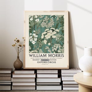 May include: A framed art print featuring a floral design in shades of white and green on a teal background. The print is labeled "WILLIAM MORRIS" with additional text about British design and location details. The print is displayed on a stack of books.