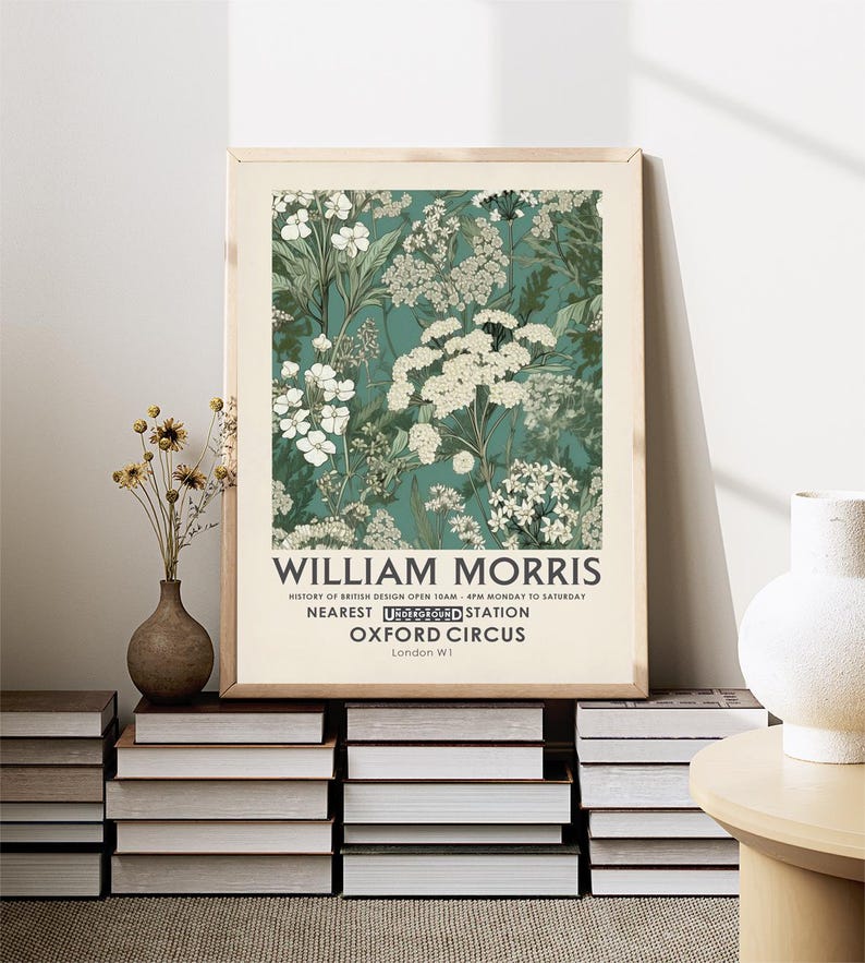May include: A framed art print featuring a floral design in shades of white and green on a teal background. The print is labeled "WILLIAM MORRIS" with additional text about British design and location details. The print is displayed on a stack of books.