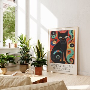 May include: A black cat with yellow eyes sits on a colourful background with flowers and swirls. The poster is framed in gold and has the text "Gustav Klimt Mus&eacute;e d'Orsay" at the bottom.