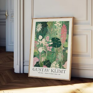 May include: A framed art print featuring a botanical illustration in shades of green, pink, and white. The artwork, attributed to Gustav Klimt, showcases various plant species. The print is displayed in a light brown wooden frame, leaning against a white wall.