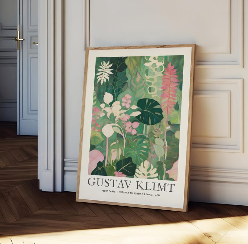 May include: A framed art print featuring a botanical illustration in shades of green, pink, and white. The artwork, attributed to Gustav Klimt, showcases various plant species. The print is displayed in a light brown wooden frame, leaning against a white wall.