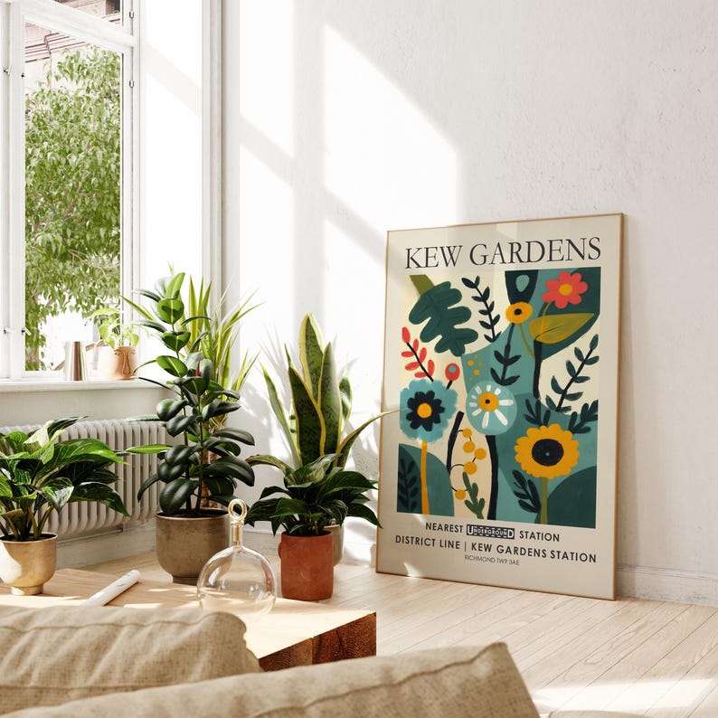 May include: A framed poster with the text "KEW GARDENS" and a colorful floral design. The poster is propped against a wall near several potted plants. The room is bright with natural light.