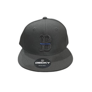 May include: Black baseball cap with a black embroidered letter "B" with a thin blue line across the centre. The cap has a black bill and a black "DECKY ORIGINAL" label on the front.