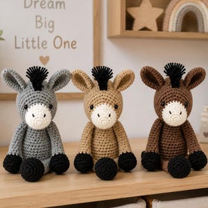 May include: Three handmade crochet donkey toys sitting in a row. The left donkey is gray, the middle is tan, and the right is dark brown. Each donkey features a white muzzle, black hooves, black ears, and a black tuft of hair on its head.