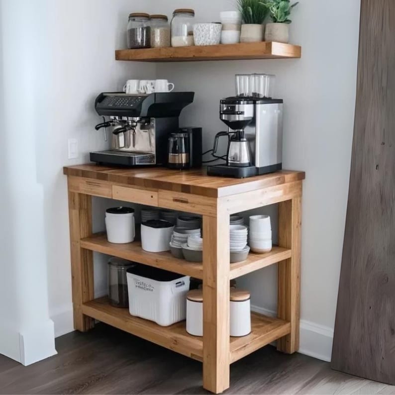 May include: A wooden coffee station featuring a black espresso machine and a silver coffee maker on the countertop. The unit includes two open shelves below holding white ceramic canisters, bowls, and mugs. A matching wooden shelf is mounted above.