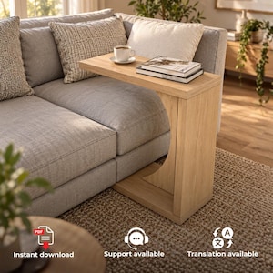 May include: A light oak wood C-shaped side table featuring a flat rectangular top surface. The table is positioned over a grey upholstered sofa and holds a white ceramic coffee cup on a saucer and a stack of two books with white covers.
