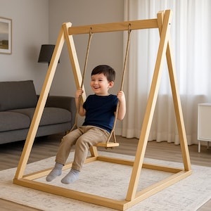 Op de afbeelding: Een houten schommel met een lachend kind. De schommel is gemaakt van lichtgekleurd hout en heeft een rechthoekige basis en A-vormige steunen. De schommelstoel is opgehangen aan een touw. Het kind draagt een marineblauw shirt en een kaki broek.