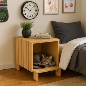 May include: A wooden bedside table with a cat bed built in. A tabby cat is asleep on a grey cushion inside the table. A black clock and a small potted plant sit on top. The table is next to a bed with grey bedding.