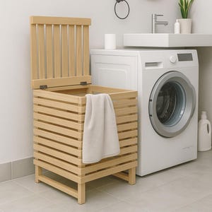May include: A light-coloured wooden laundry hamper with a slatted design and a hinged lid. A beige towel hangs over the edge. The hamper is in a bathroom setting, next to a white washing machine.