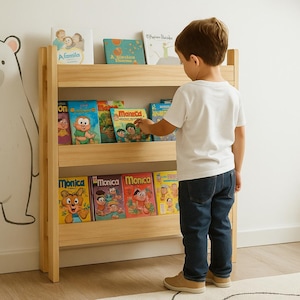 以下が含まれることがあります： カラフルな子供向けの本でいっぱいの明るい色の木製本棚。本にはさまざまなイラストとタイトルがあります。若い人が真ん中の棚の本に手を伸ばしています。本棚は、クマのイラストが描かれた白い壁に立てかけられています。