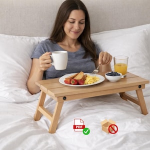 May include: A light brown wooden breakfast tray with folding legs, holding a plate of food, a bowl of blueberries, and a glass of orange juice. A woman is holding a white mug. The tray is on a bed with white linens.