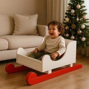 Könnte beinhalten: Ein weißer Holzspielzeugschlitten mit roten Kufen. Der Schlitten hat eine geschwungene Vorder- und Rückseite und einen roten Sitz. Der Schlitten ist für ein Kind zum Sitzen konzipiert. Ein Weihnachtsbaum ist im Hintergrund.