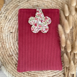 Peut inclure: Une pochette rectangulaire en velours côtelé magenta avec un fermoir floral. Le fermoir est en forme de fleur avec un centre blanc et un motif de petites fleurs rouges et roses sur fond blanc. La pochette est posée sur un tapis rond tressé.