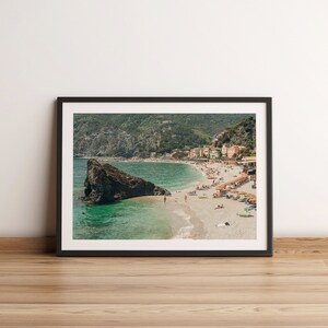 K&ouml;nnte beinhalten: Ein gerahmtes Foto einer Strandszene mit einer gro&szlig;en Felsformation im Vordergrund. Der Strand ist voller Menschen und im Hintergrund befinden sich Geb&auml;ude am Hang.