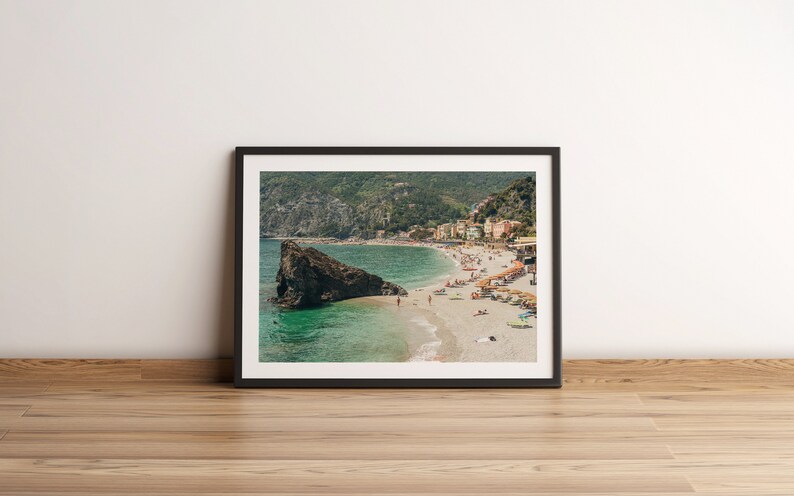 K&ouml;nnte beinhalten: Ein gerahmtes Foto einer Strandszene mit einer gro&szlig;en Felsformation im Vordergrund. Der Strand ist voller Menschen und im Hintergrund befinden sich Geb&auml;ude am Hang.