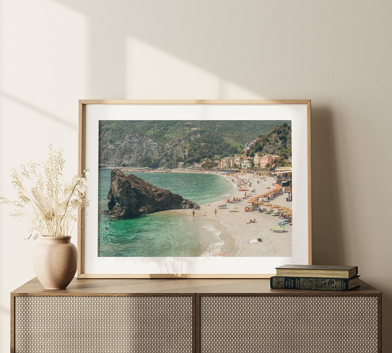 K&ouml;nnte beinhalten: Ein gerahmtes Foto einer Strandszene mit einem felsigen Vorsprung im Vordergrund, einem t&uuml;rkisfarbenen Meer und einem kleinen Ort an einem Hang im Hintergrund. Der Strand ist voller Menschen, die die Sonne und den Sand genie&szlig;en.