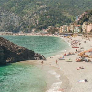 K&ouml;nnte beinhalten: Blick auf einen Strand mit einem felsigen Vorsprung im Wasser. Der Strand ist voller Menschen und Sonnenschirme. Hinter dem Strand befinden sich Geb&auml;ude am Hang.