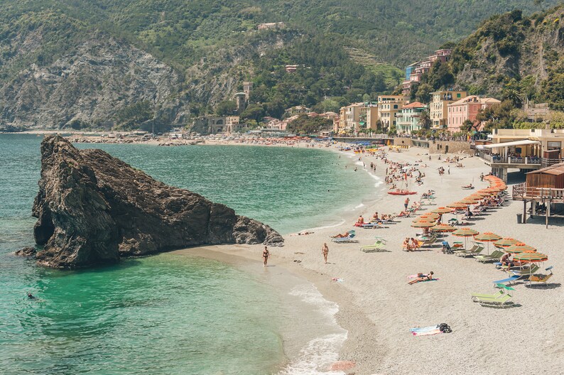 K&ouml;nnte beinhalten: Blick auf einen Strand mit einem felsigen Vorsprung im Wasser. Der Strand ist voller Menschen und Sonnenschirme. Hinter dem Strand befinden sich Geb&auml;ude am Hang.