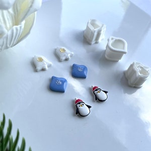 May include: A set of Christmas-themed cookie cutters, including a penguin, a snowflake, and a bell. The cutters are white plastic and are arranged on a white surface.