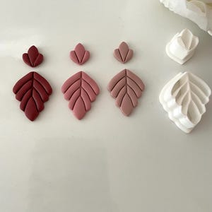 May include: Three sets of leaf-shaped fondant cutters in various sizes. The cutters are made of white plastic and are shown next to three fondant leaves in shades of pink and red.
