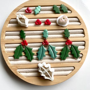 May include: A set of three pairs of green and red holly leaf earrings with white holly leaf cookie cutters on a wooden background.