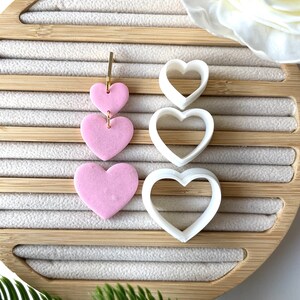 May include: Three pink heart-shaped polymer clay earrings with a gold jump ring hanging from the top heart. The earrings are displayed next to three white heart-shaped cookie cutters.