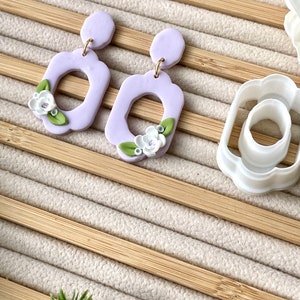 May include: A pair of lavender polymer clay earrings with a floral design. The earrings are shaped like a rectangle with a rounded top and bottom. The floral design features white flowers and green leaves.