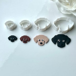 May include: A collection of four white plastic dog-shaped cookie cutters, alongside matching dog face decorations in black, brown, and tan. The dog faces have white eyes and are arranged in a row.