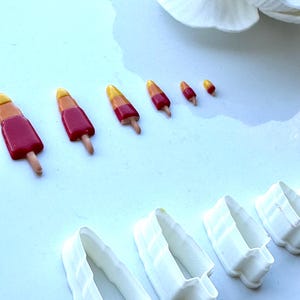 May include: Five different sized cookie cutters shaped like popsicles with a red, yellow, and orange colour scheme. The cutters are made of white plastic and are arranged in order of size from largest to smallest.