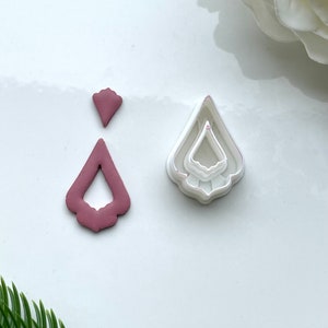 May include: A set of three white plastic cookie cutters in the shape of a teardrop with scalloped edges. The cutters are layered on top of each other, with the smallest cutter on top. A pink clay teardrop shape is shown next to the cutters.