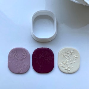 May include: Three oval-shaped polymer clay pieces with different floral designs embossed on them. The designs are a single rose, a cluster of small flowers, and a cluster of larger flowers. The clay pieces are in shades of purple, burgundy, and white.