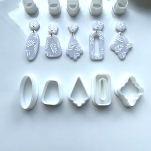 May include: A set of 10 white polymer clay earring cutters, including a variety of shapes, such as oval, rectangle, triangle, and leaf. The cutters are arranged in two rows on a white surface.