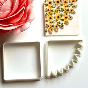 May include: A square craft piece featuring a sunflower design, a white square, and a white corner-shaped cutter. A large pink rose is in the upper left. The craft has yellow sunflowers with green leaves.