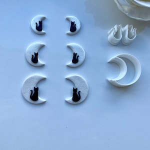 May include: Six white fondant crescent moon shapes with black cat silhouettes on a white surface. There are also two white plastic crescent moon cookie cutters and two white plastic flower cookie cutters.