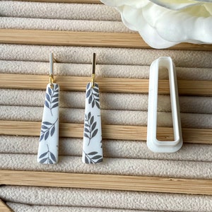 May include: A pair of dangle earrings with a white background and black leaf pattern. The earrings are made of ceramic and have gold-coloured hooks.