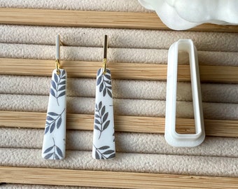 May include: A pair of dangle earrings with a white background and black leaf pattern. The earrings are made of ceramic and have gold-colored hooks.