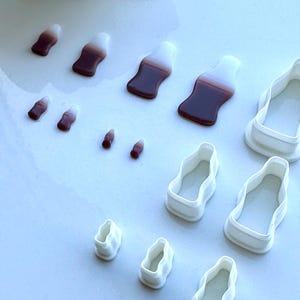 May include: A collection of small, brown and white soda bottle-shaped sweets and white plastic cookie cutters. The sweets have a gradient effect from white to brown. The cookie cutters are in the shape of the soda bottles.