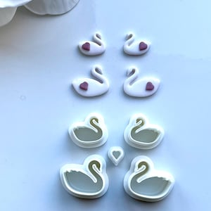 May include: Set of six white plastic swan cookie cutters. Four of the cutters feature a small pink heart on the swan's chest. Two of the cutters are larger and do not have hearts.