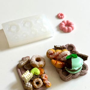 May include: Miniature food models including donuts, pastries, and a macaron, presented on small brown trays. A clear silicone mould with donut shapes is also visible. The food items are in various colours, including pink, brown, and white.