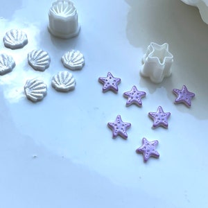 May include: White plastic cookie cutters shaped like a seashell and a star, with purple sugar paste seashells and star-shaped cookies.