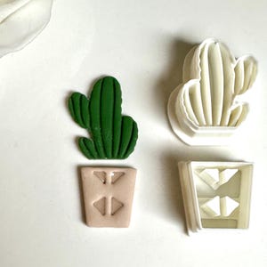May include: A green cactus in a tan pot, and a white cactus-shaped cookie cutter, and a white pot-shaped cookie cutter. The cactus has a detailed design. The pot has a geometric design. The items are on a white surface.