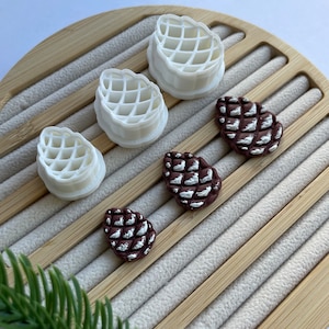 May include: Five pine cone shaped ornaments, two are white and three are brown with silver glitter. The ornaments are on a wooden surface.