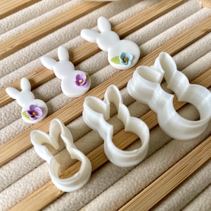 May include: Five white plastic bunny cookie cutters of varying sizes. The cutters are on a wooden surface with a light brown background.