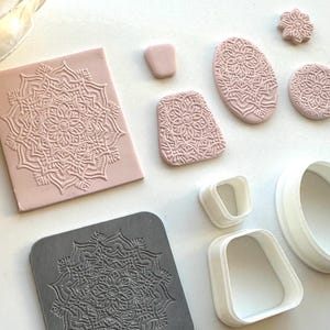 May include: A selection of pink and grey mandala-patterned clay stamps and cutters. The stamps are in various shapes, including rectangular, oval, and circular. White plastic cutters are also present, in square and rounded shapes. The items are arranged on a white surface.