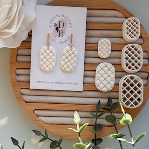 May include: White and gold earrings with a quilted pattern, presented on a wooden tray. The earrings are rectangular with gold detailing. The tray also holds matching earring moulds. A card with the text "DAVINIA BROOKE CLAY DESIGNS" is visible.