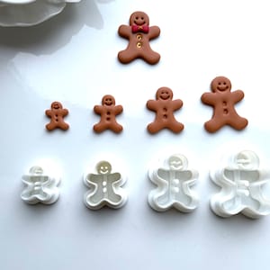 May include: Gingerbread man cookie cutters and decorated cookies in various sizes. The cookies are brown with white icing details and a red bow tie. The cutters are white plastic.