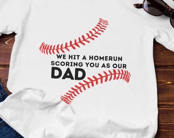 May include: White t-shirt with the text "WE HIT A HOMERUN SCORING YOU AS OUR DAD" in black and red. The design includes baseball stitching. Sunglasses and a small succulent plant are also visible.