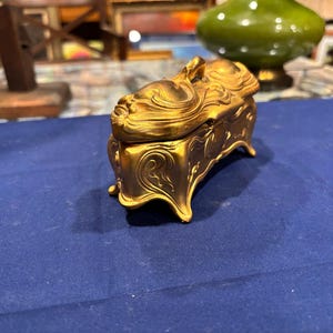 May include: A gold-colored jewelry box with a detailed, ornate design. The box has a curved lid with a sculpted figure on top. The box sits on four curved feet. The background is a blue surface.